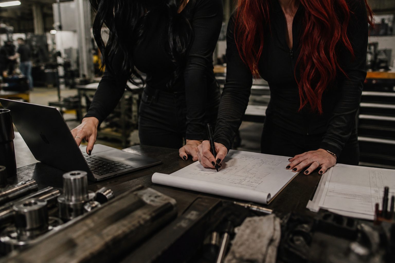 Hands marking up paper blueprints beside a laptop on a machine shop bench